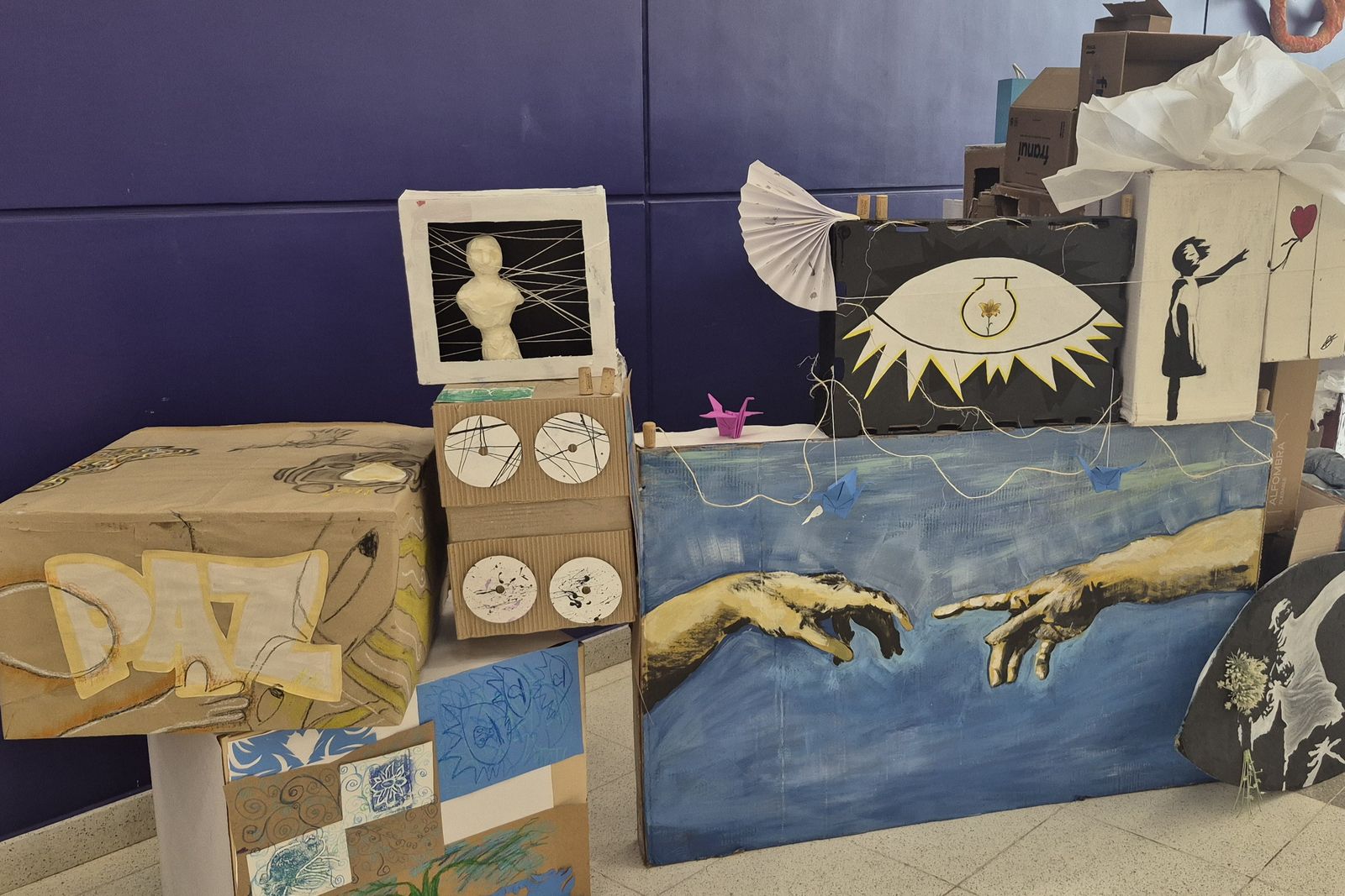 Obras de la Expo Arte 2026: una instalación de cartón con dos manos pintadas sobre fondo azul, junto a otras piezas de alumnos