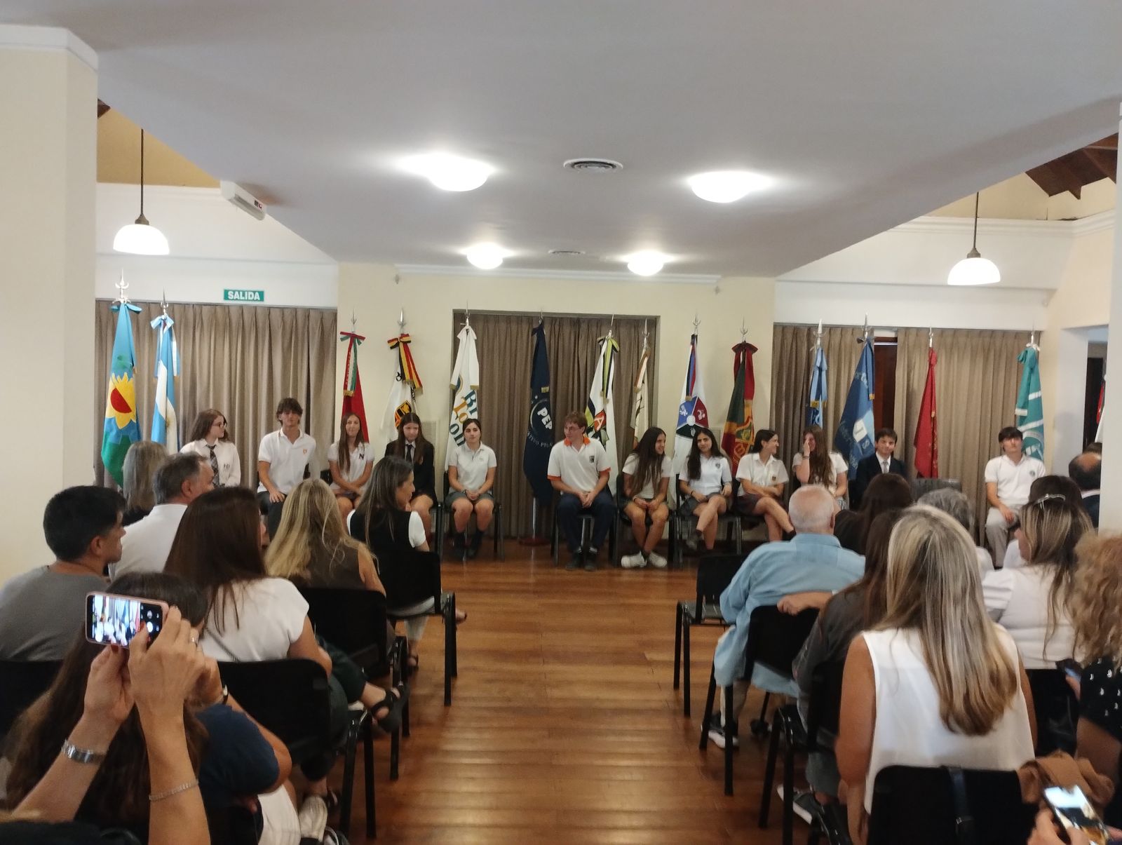 Ceremonia de la Medalla al Mérito: alumnos sentados al frente, rodeados de banderas, y las familias en el público